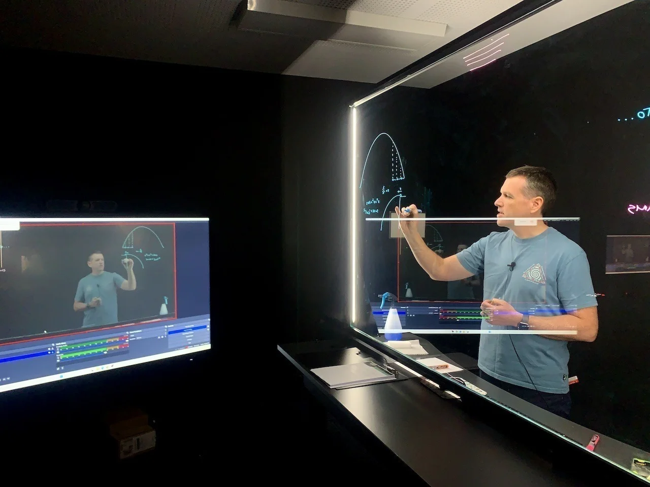 An instructor draws on an illuminated lightboard while a nearby monitor displays the recorded explanation.